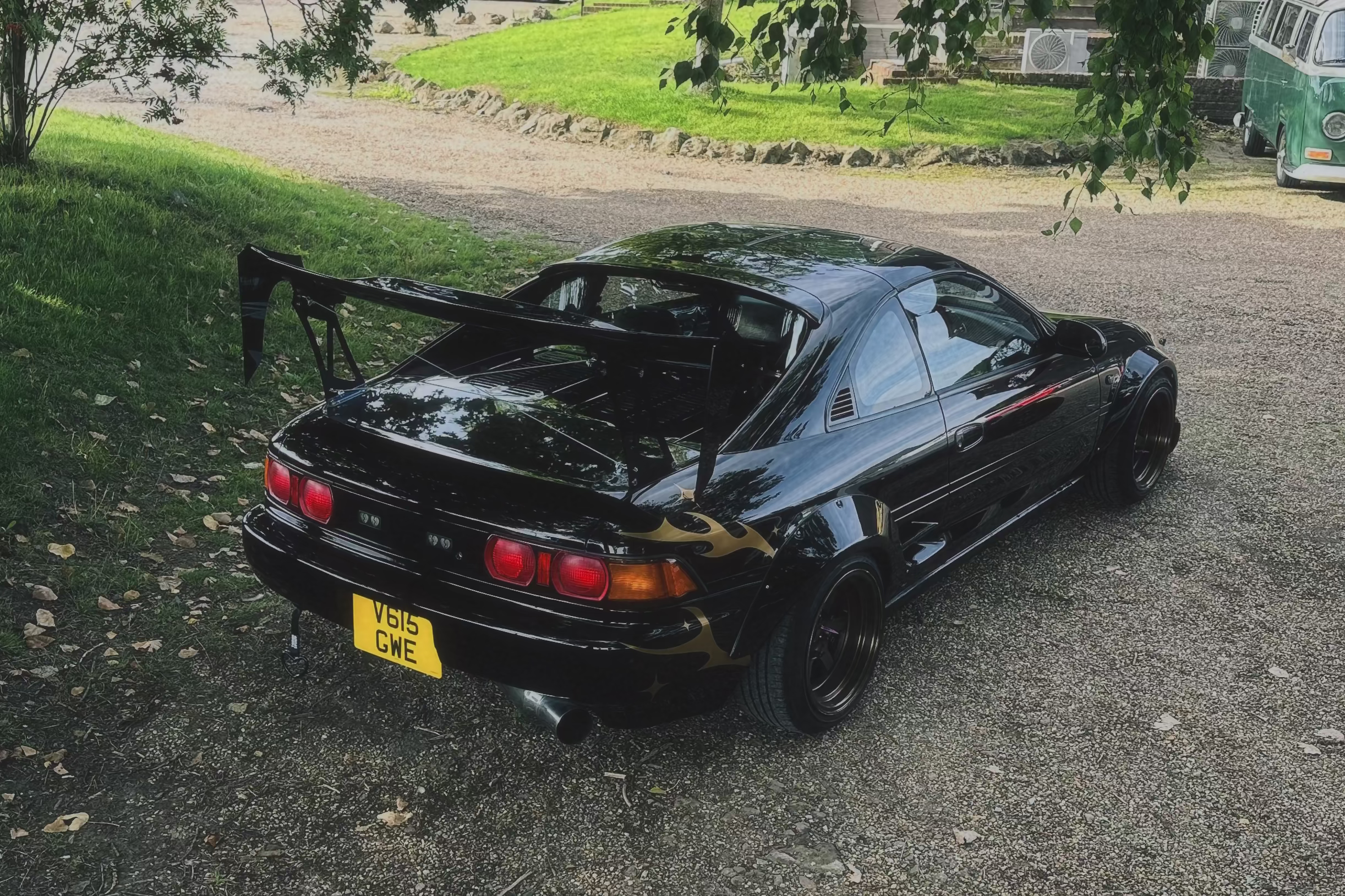 A black widebody Toyota MR2 with a large rear wing is parked on a gravel driveway in a grassy outdoor area with trees and a house in the background.